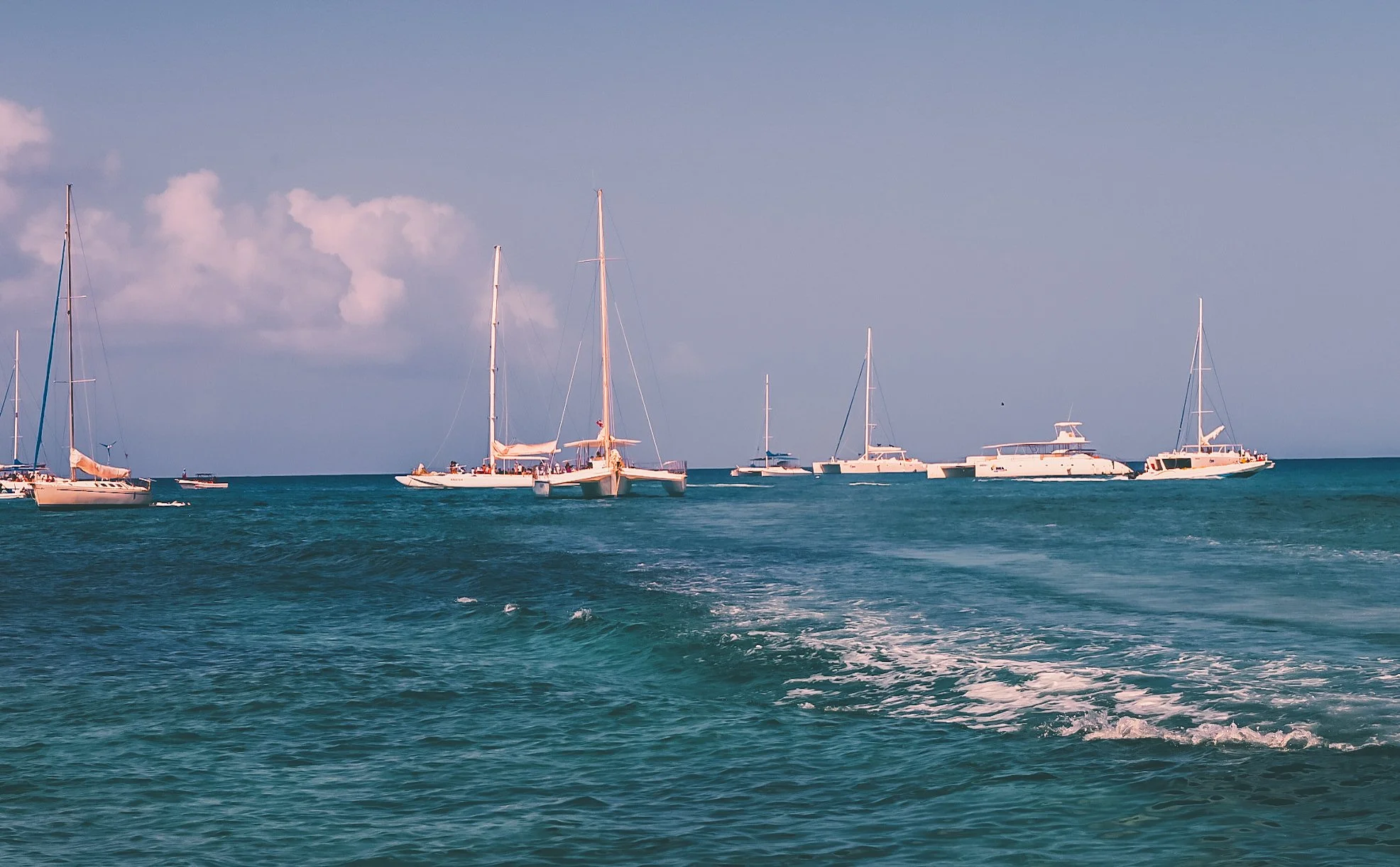 Several sailboats anchored in calm ocean water under a partly cloudy sky.