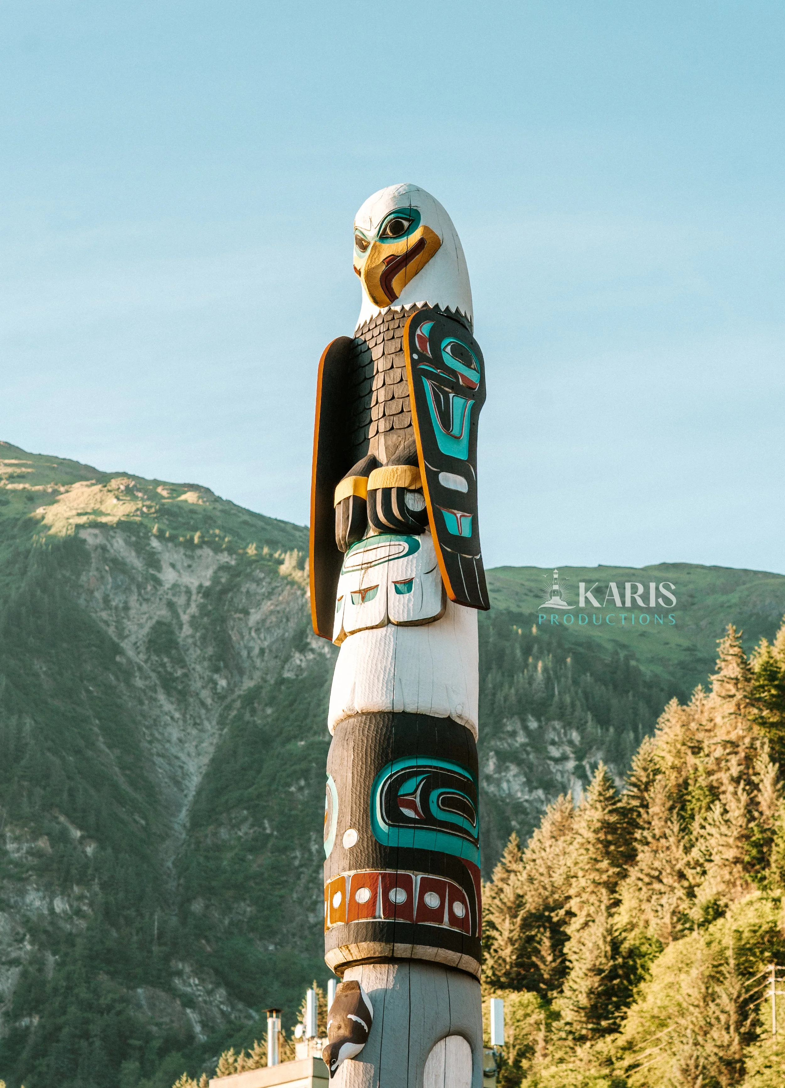 A tall carved totem pole painted with indigenous designs and animals, set against a backdrop of green mountains and a clear blue sky. Juneau, Alaska. Karis Productions copyright 2025