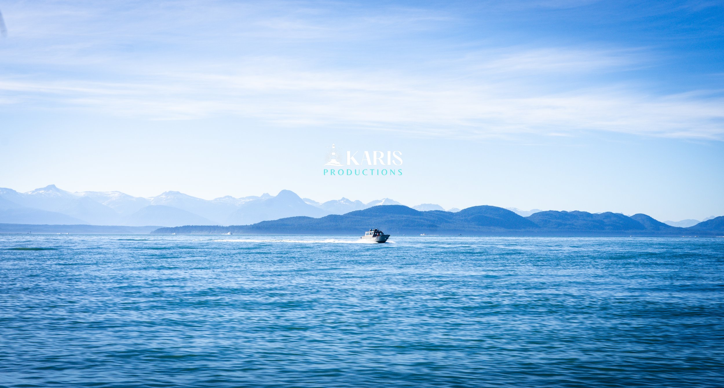 A boat sailing on calm blue waters with mountains in the background and a partly cloudy sky above. Juneau Alaska. Karis Productions copyright 2025