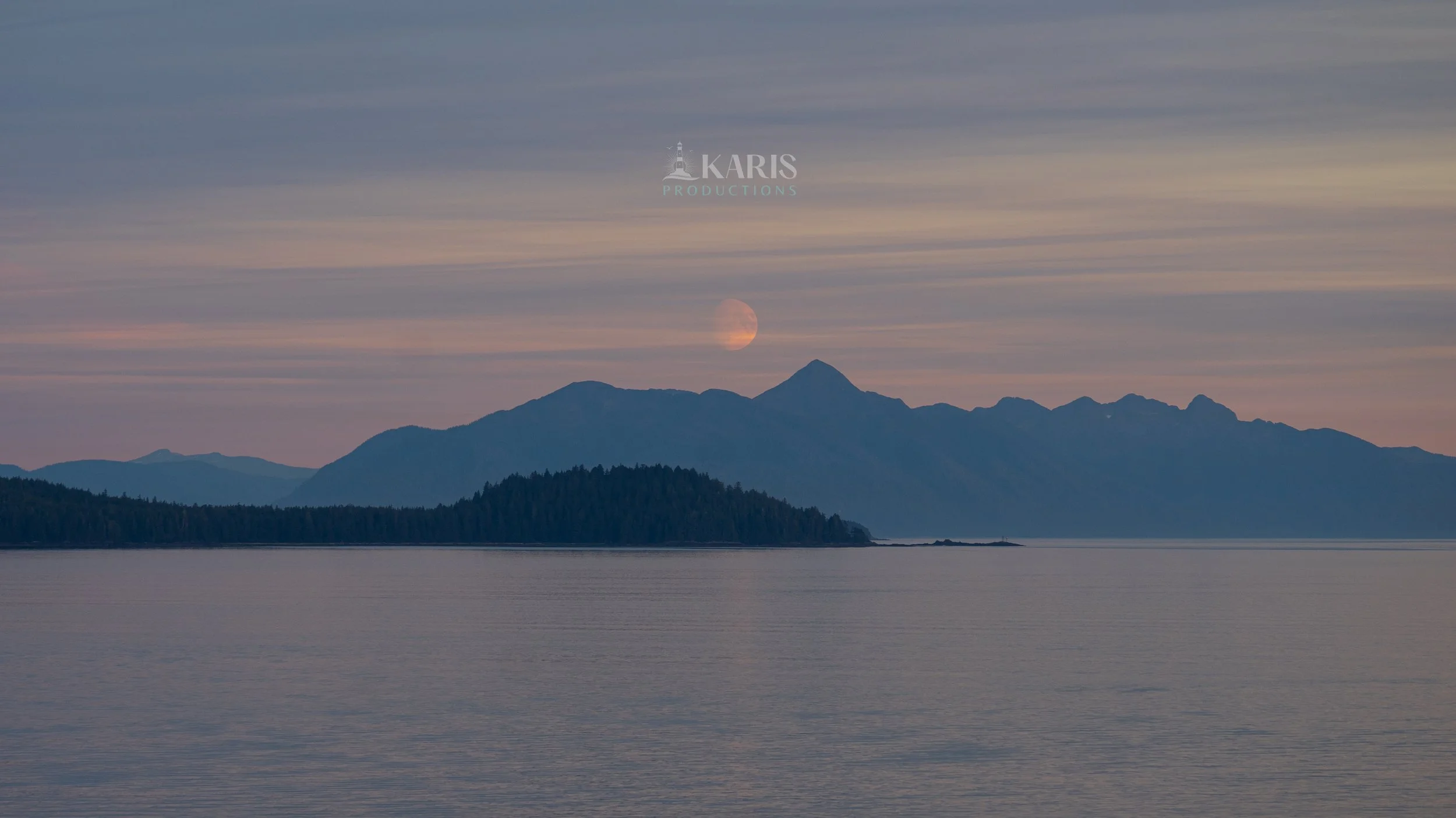 Scenic view of mountains with the moon rising behind them, reflected on a large body of water in the foreground, during dusk or dawn.