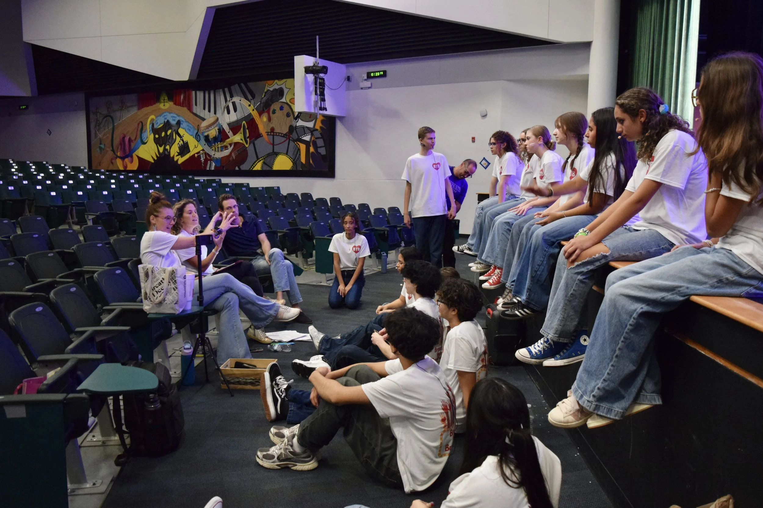 Group of young people sitting and standing in a theater or auditorium, some are talking and others are taking photos. The auditorium has rows of empty seats and colorful murals on the walls.