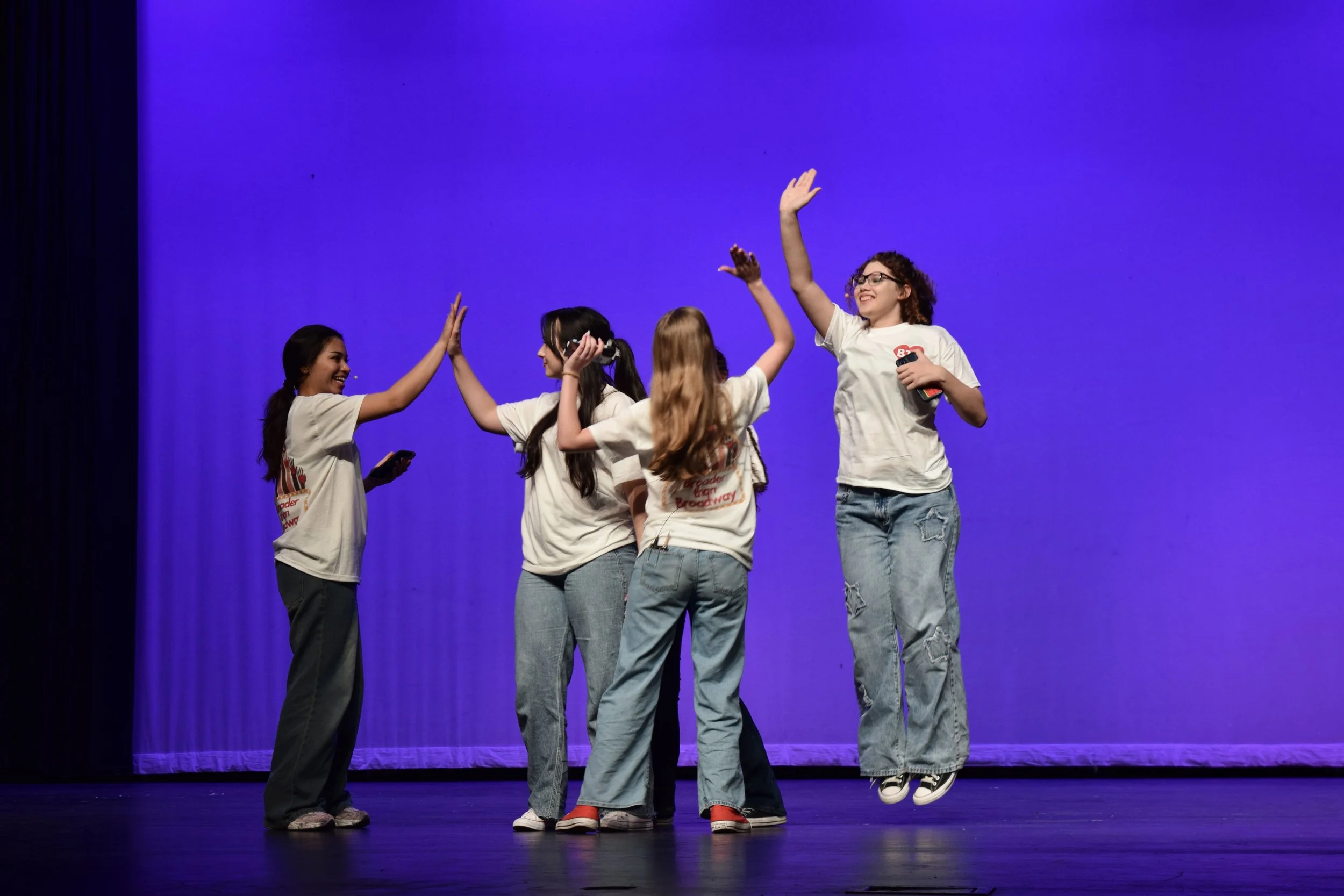 Group of young people sitting and standing in a theater or auditorium, some are talking and others are taking photos. The auditorium has rows of empty seats and colorful murals on the walls.
