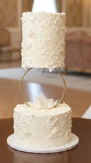 Elegant double-tiered white wedding cake with floral decorating set to the bridal dress lace and a unique gold ring support on a cake stand. Bespoke luxury wedding cakes in Calgary by Magnolia Couture Cakery.