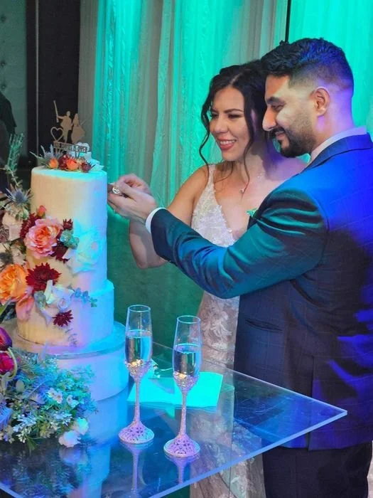 Three-tier wedding cake with fresh floral decorations and a custom topper, accompanied by a dessert menu on a glass table. for a luxury wedding in calgary
