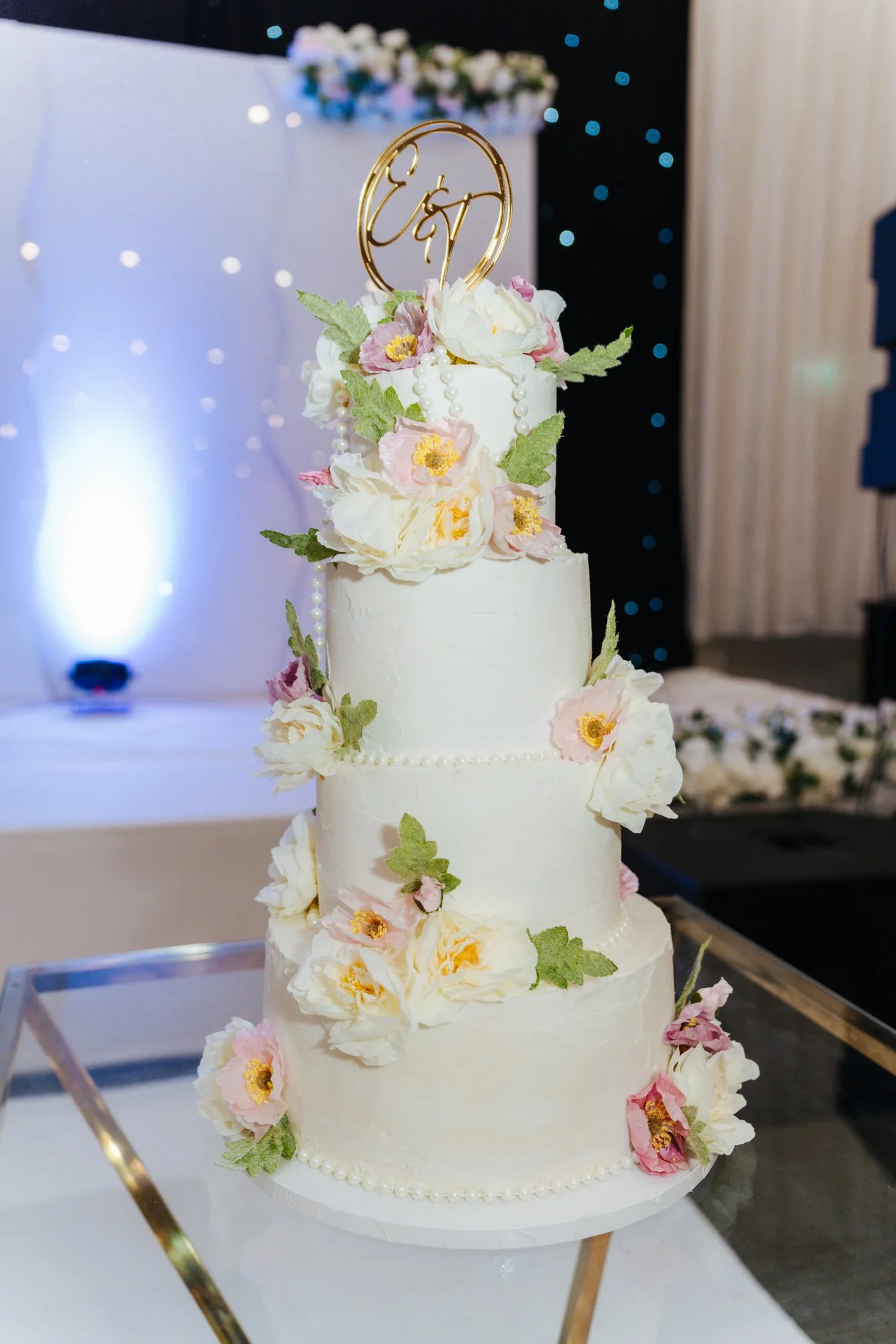 This elegant four-tier wedding cake features a smooth white finish accented with delicate pearl borders and a cascading arrangement of flowers. Large ivory blooms, soft blush blossoms, and textured green sugar leaves are individually handcrafted and 