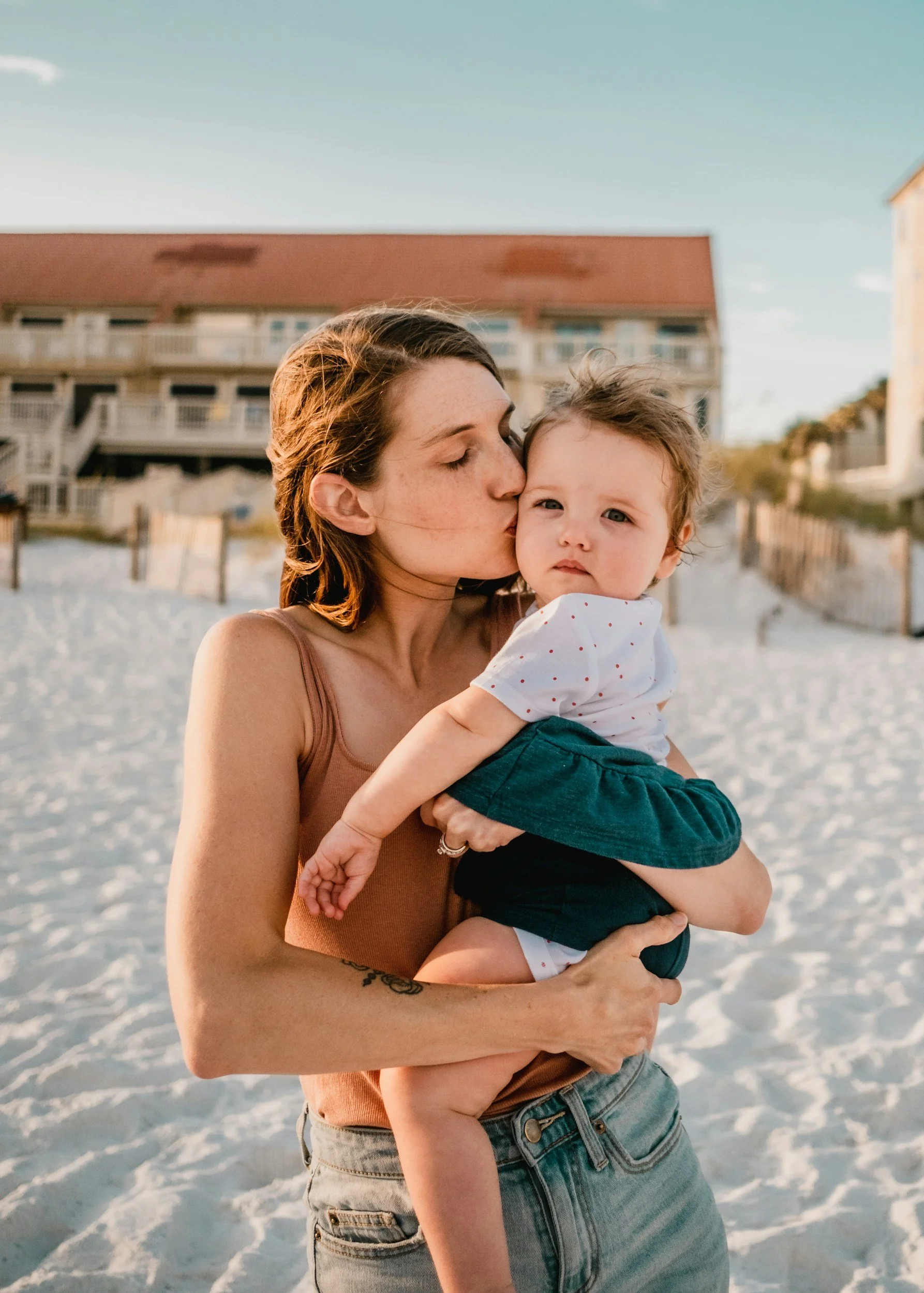 A woman with brown hair kissing a young boy on the cheek at the beach. The woman is wearing a sleeveless top, and the boy is wearing a white shirt with red dots and dark shorts. They are standing on sand with buildings and a wooden fence in the background, under a clear sky.