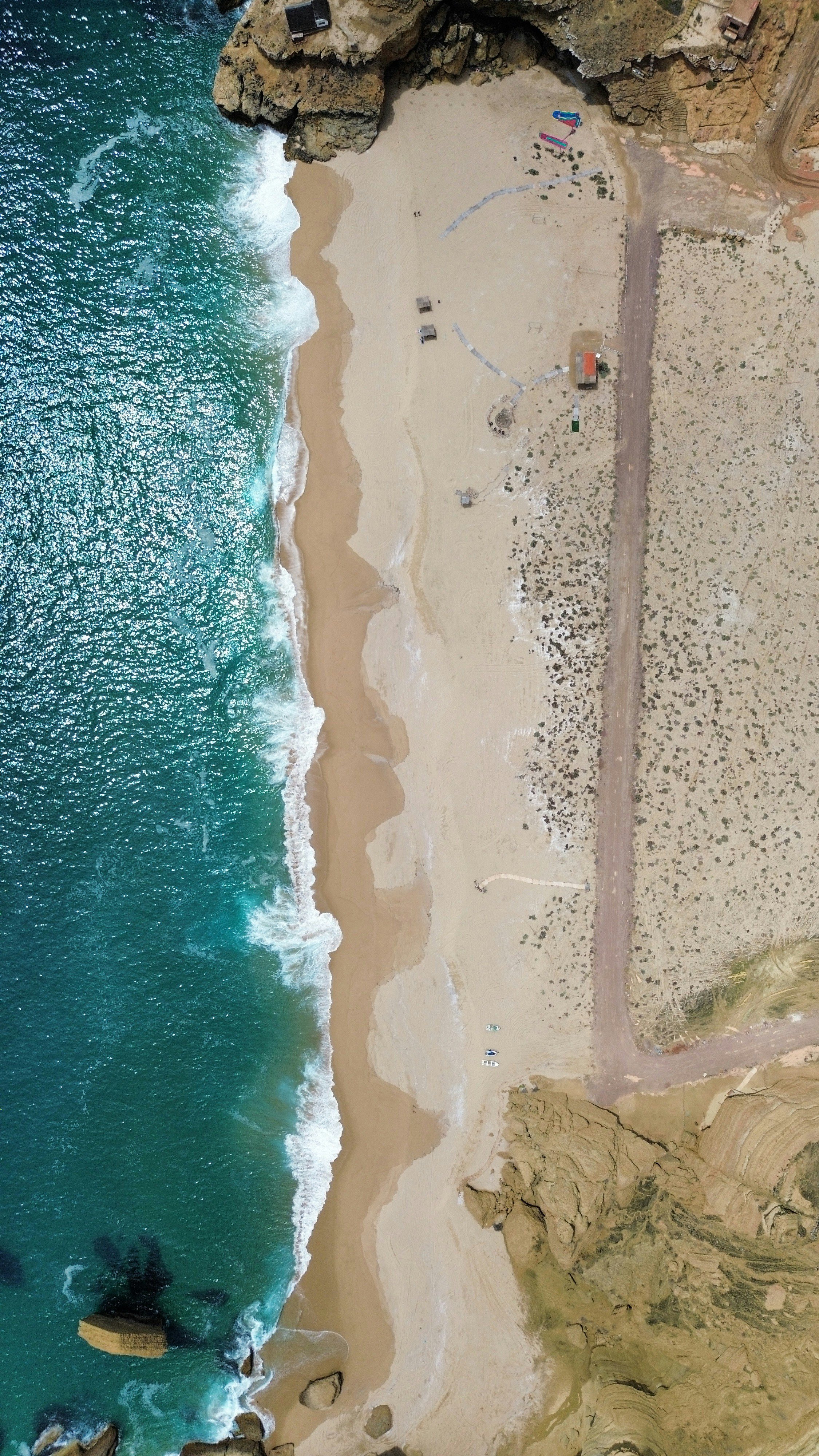 An aerial view of a beach with waves crashing on the sandy shore, rocky cliffs on one side, and some structures and pathways on the land area.