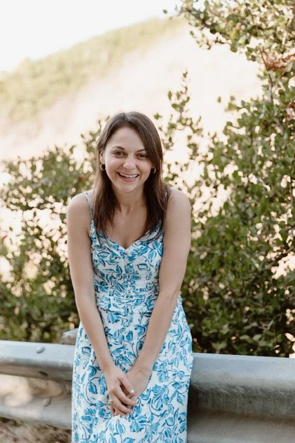 Morgan Abraham, LMFT in a blue and white floral dress, standing outdoors near a guardrail, smiling at the camera with greenery and trees in the background.