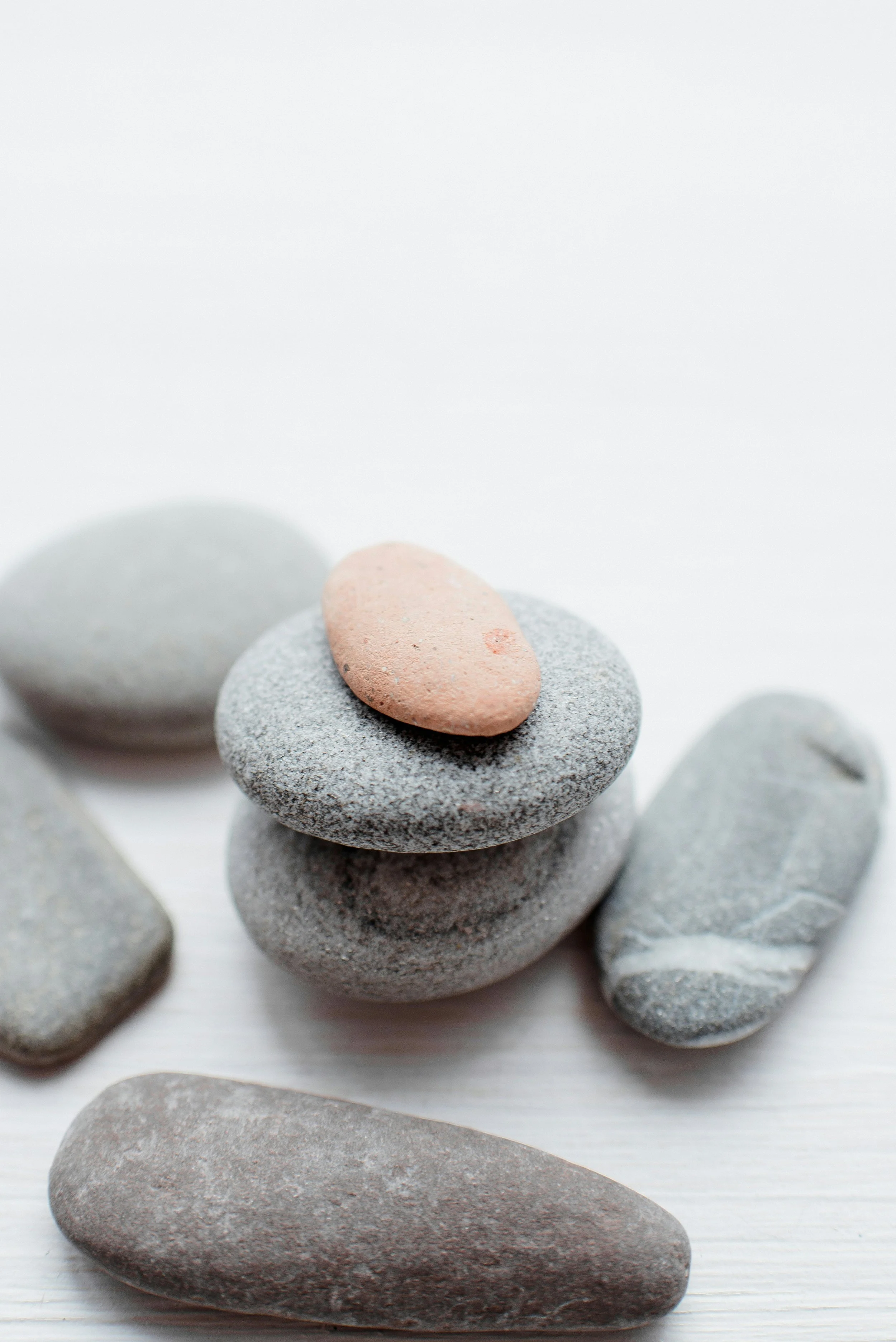 A stack of smooth, rounded stones of various sizes and colors, with the top stone being a small, pinkish pebble. The stones are arranged on a light surface with few other stones visible nearby.