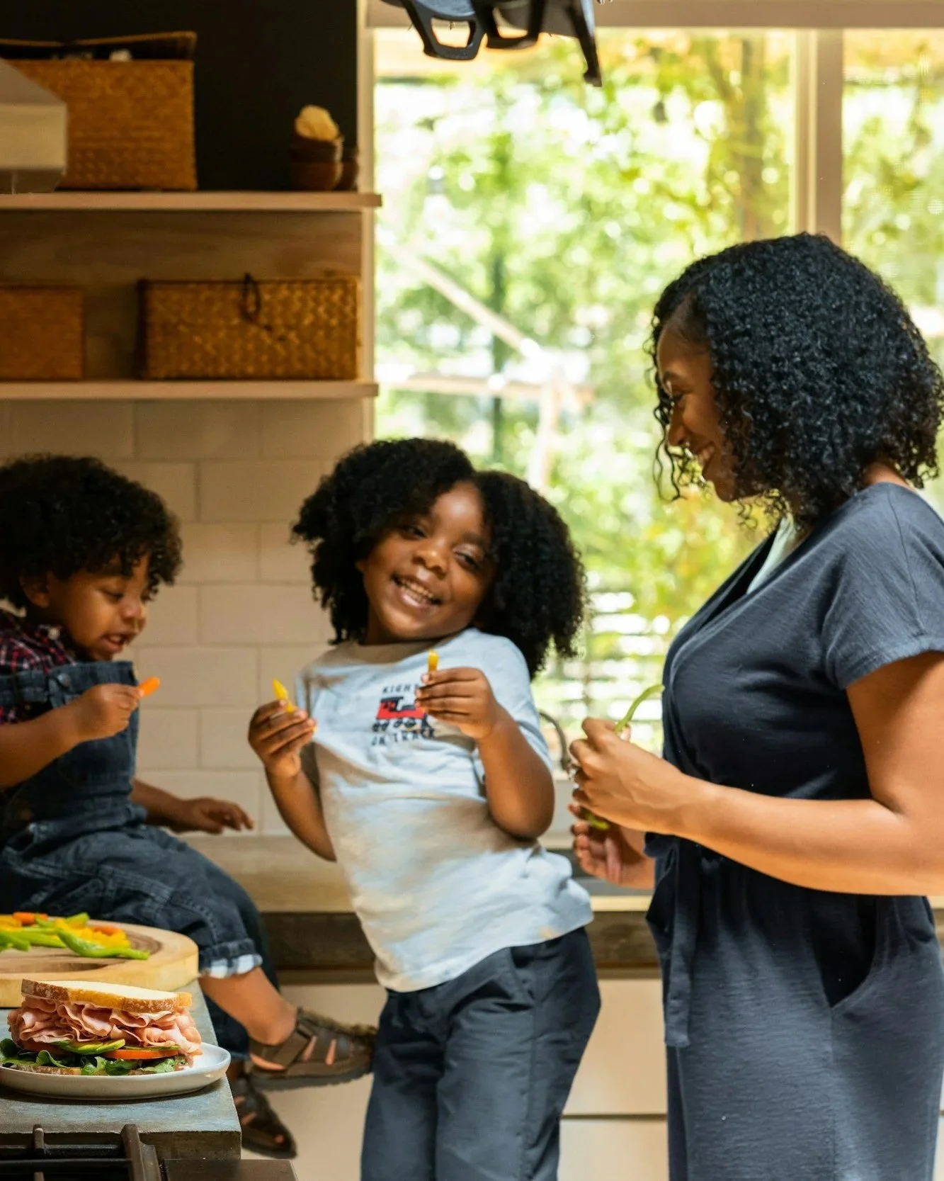 A woman and two children in a kitchen, smiling and holding yellow and orange peppers. The girl in the middle is smiling at the camera, while the boy on the left is focused on his pepper. The kitchen has wooden shelves with woven baskets and a window with greenery outside.