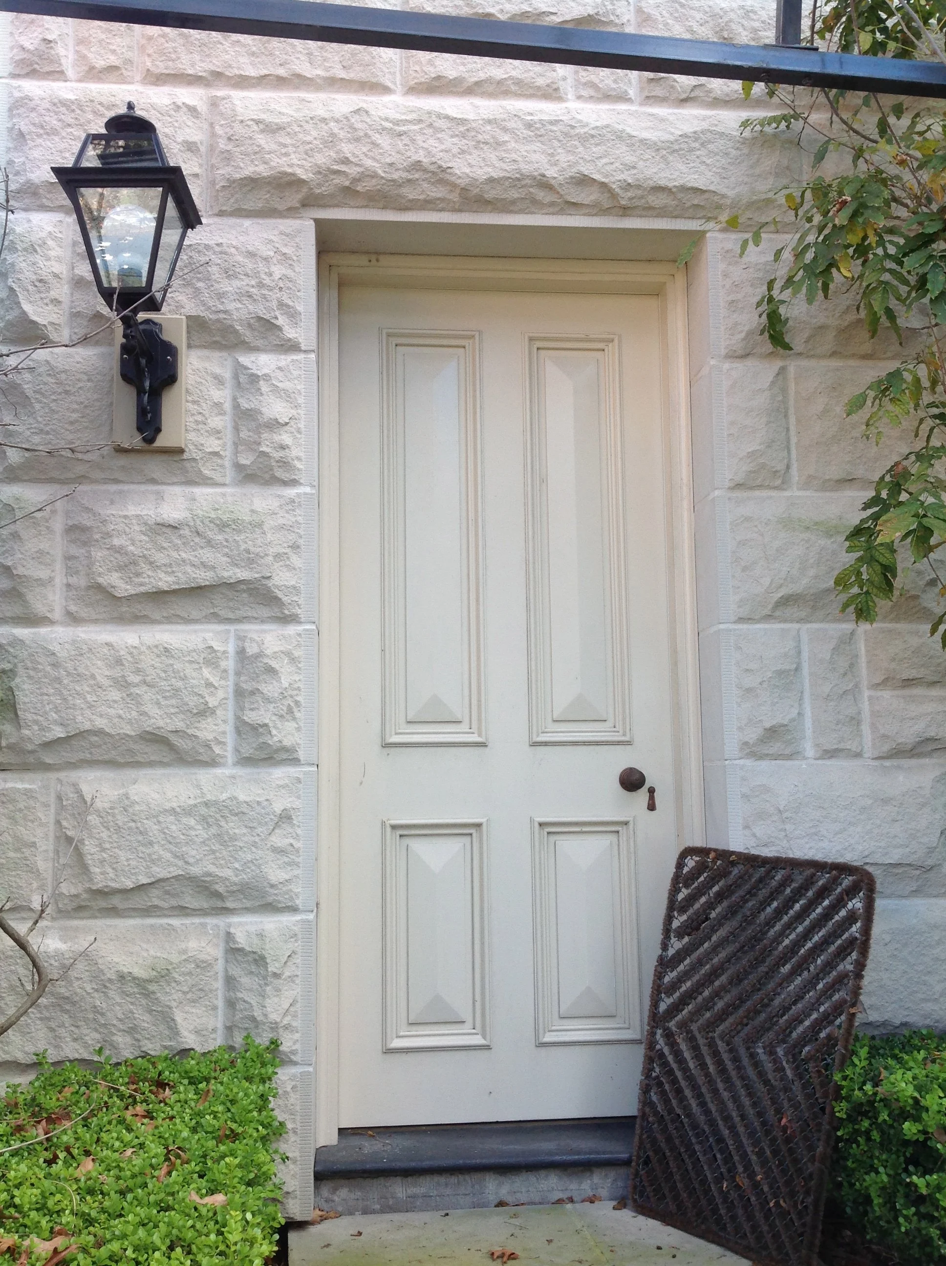 White door with ornate panels, a black lantern-style light on stone wall, bush and metal grate on the ground.