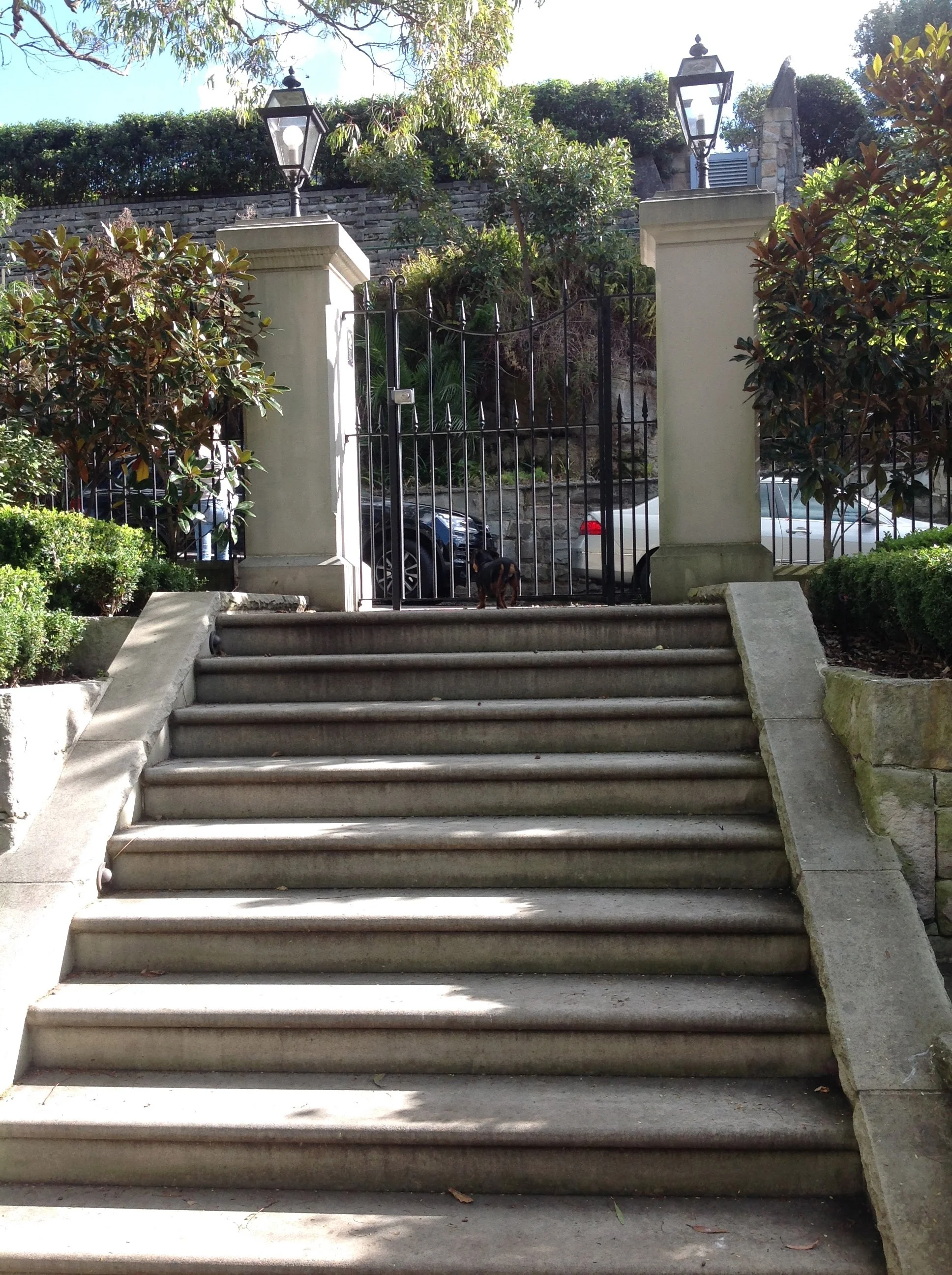 Stone staircase leading up to a black wrought-iron gate, flanked by two stone pillars with lanterns. Trees and bushes surround the area, and parked cars are visible behind the gate.