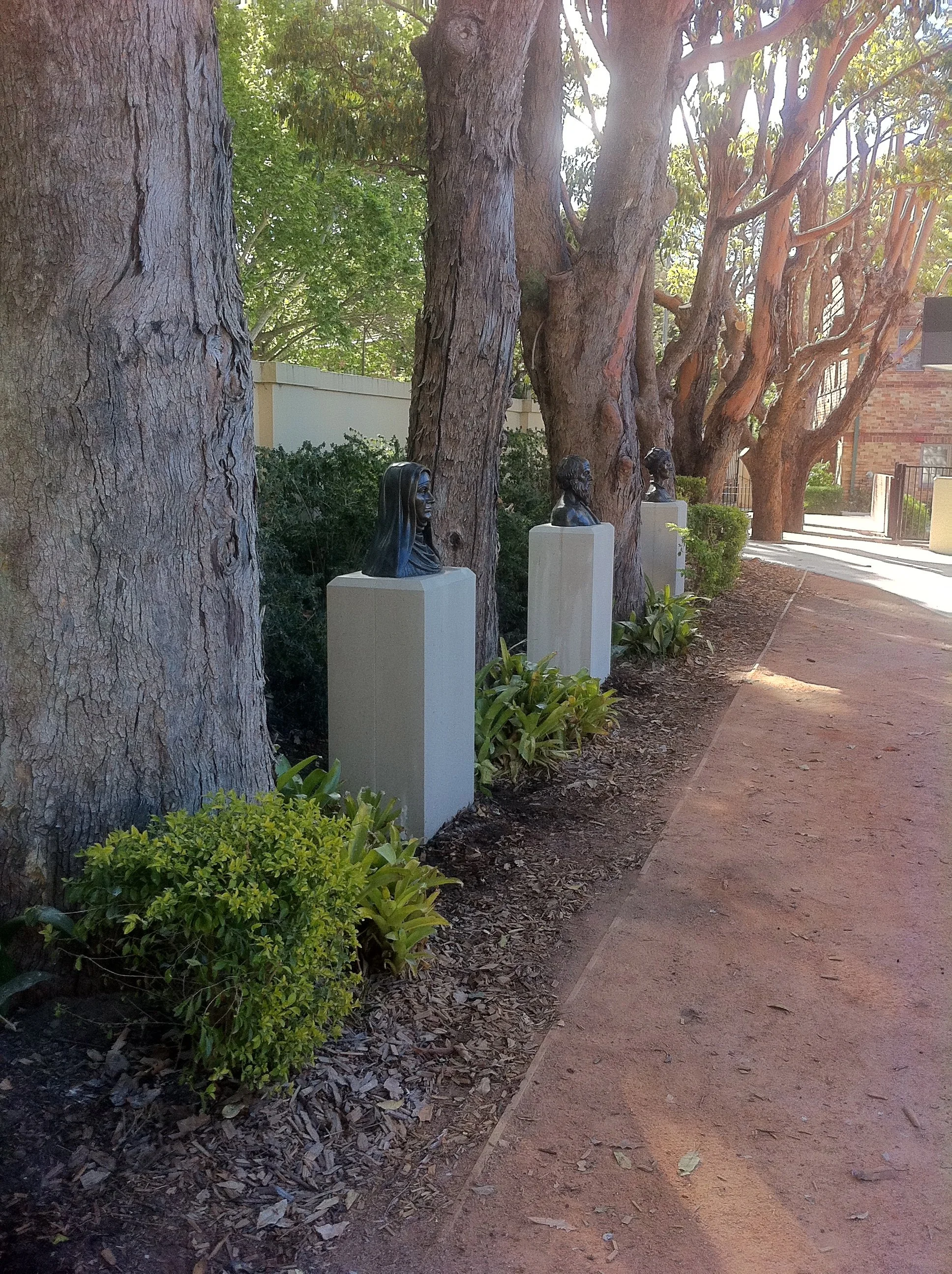 Bust sculptures on pedestals in a garden pathway lined with trees.