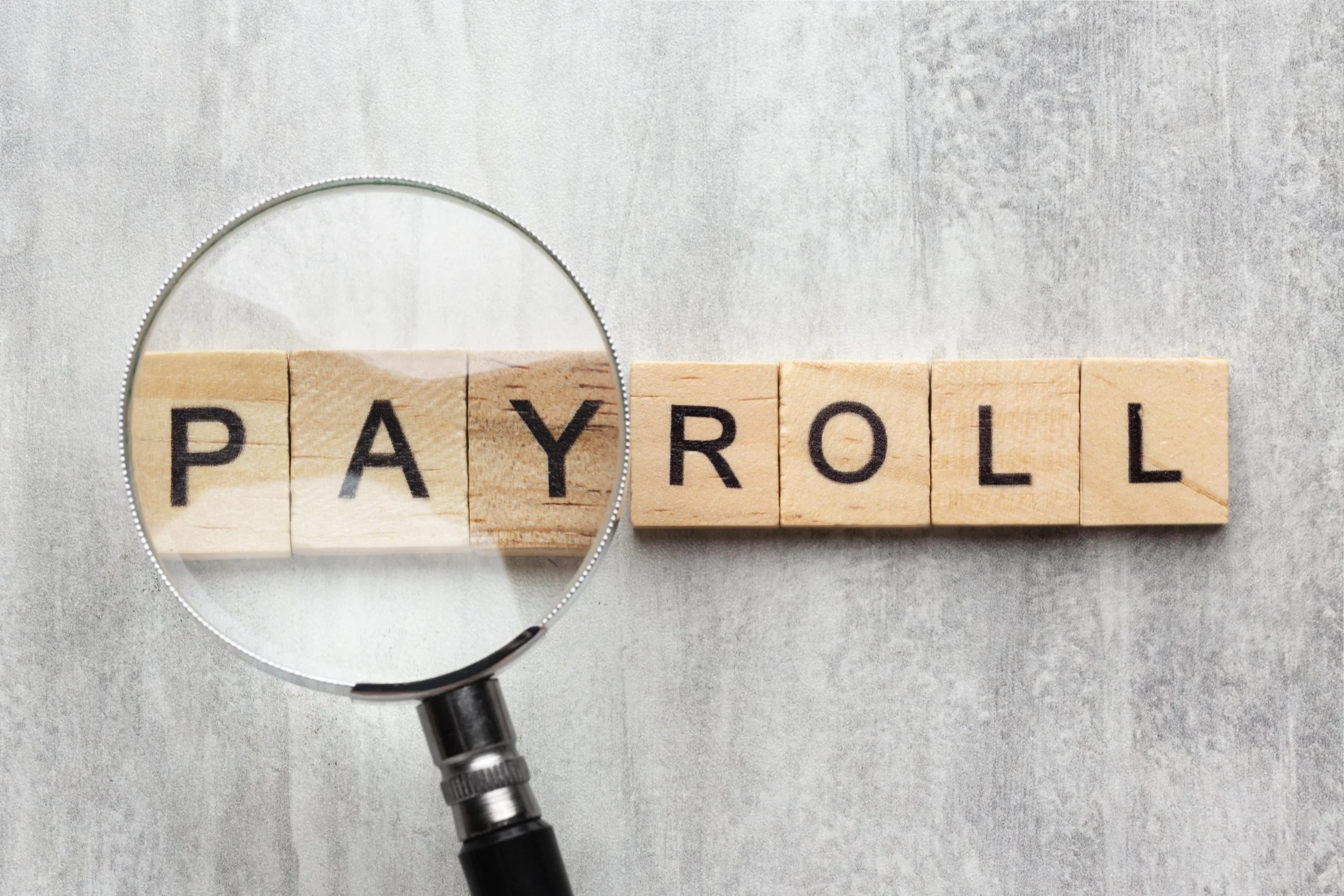 Close-up of wooden letter tiles spelling 'PAYROLL' with a magnifying glass examining part of the word, placed on a gray surface.