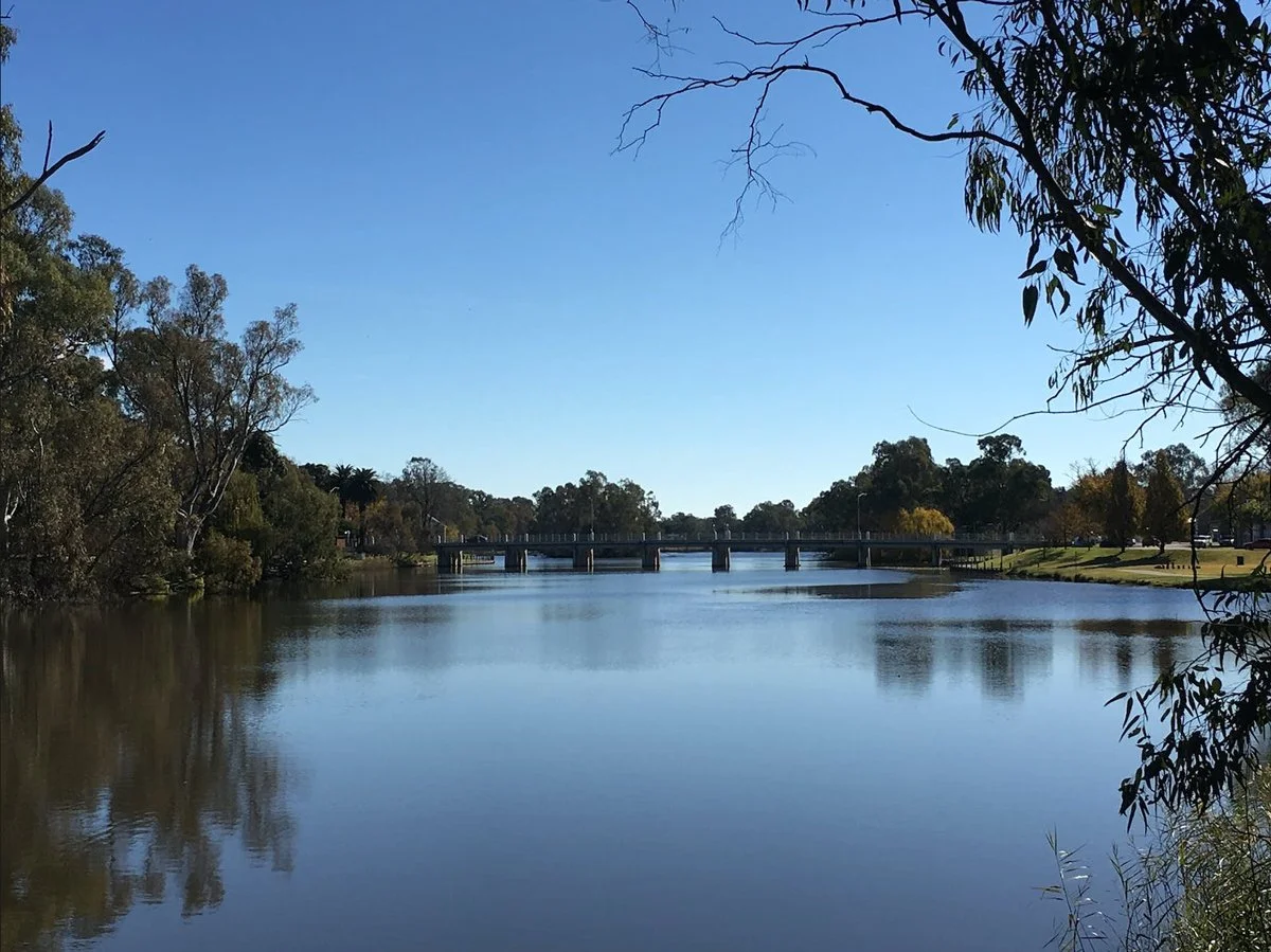 A peaceful river scene with calm water reflecting trees on both sides and a clear blue sky above, with a bridge across the river in the distance.