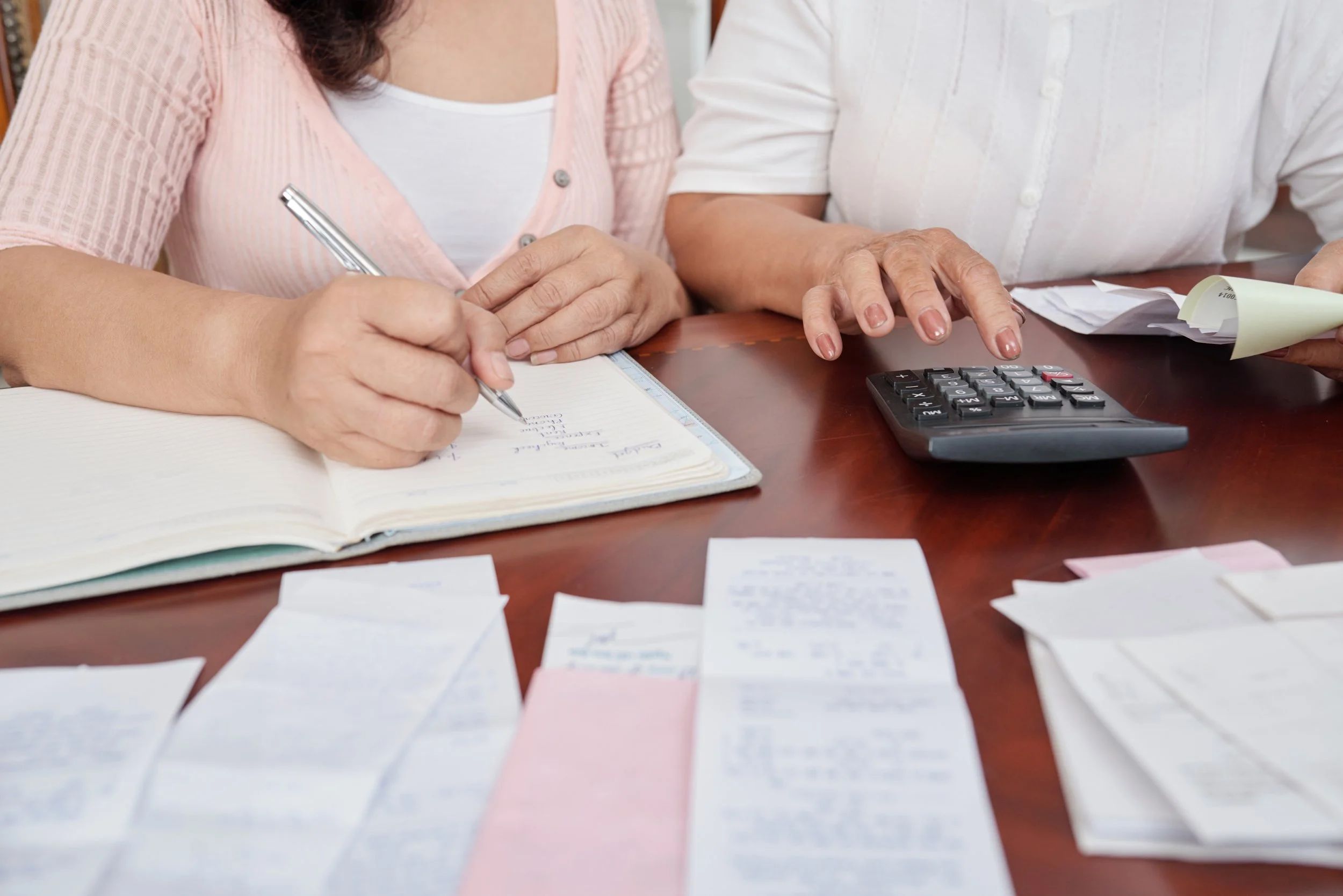 Two women working at a desk with papers, receipts, a calculator, and a pen.