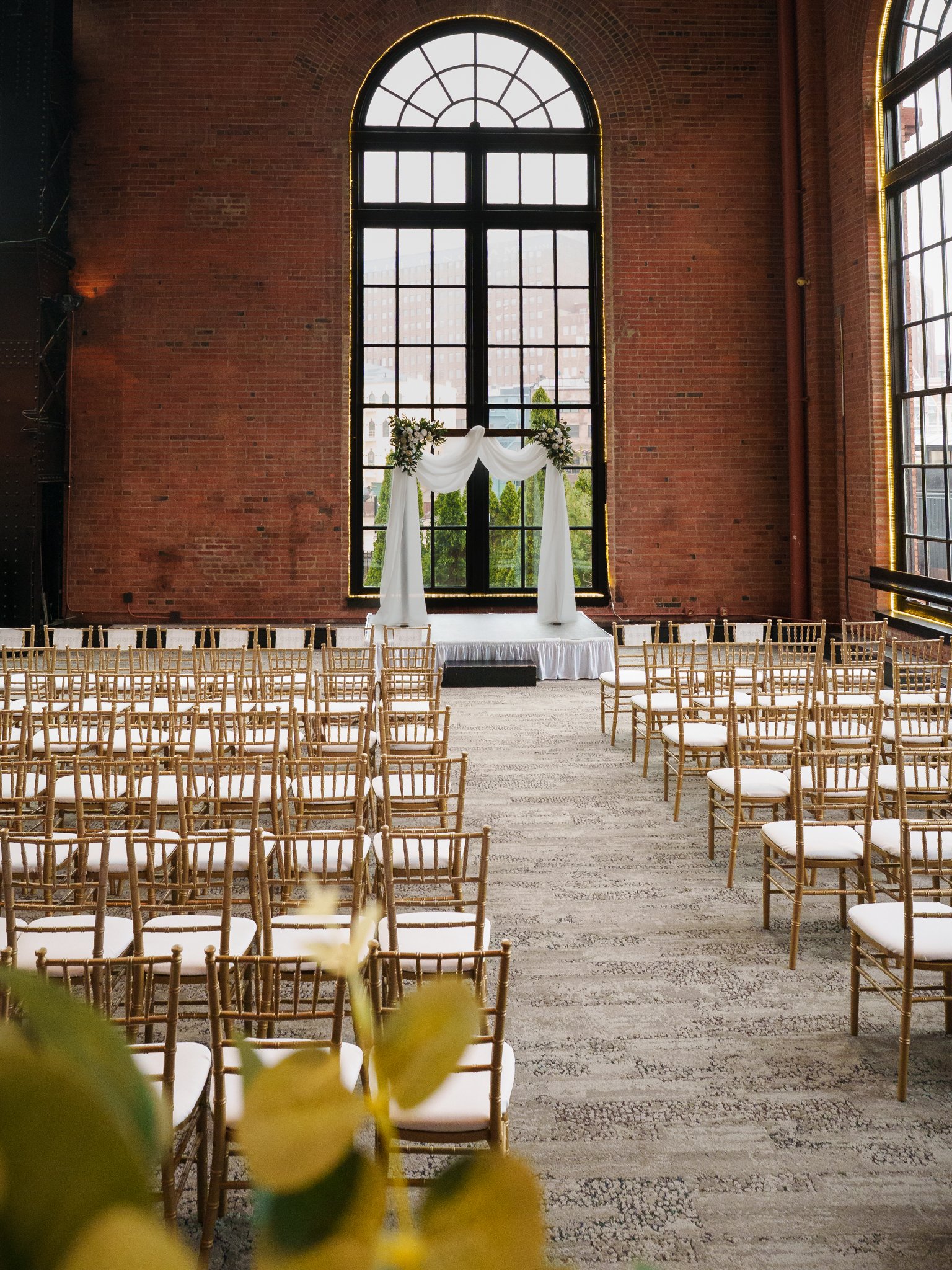 Indoor wedding ceremony setup with rows of gold chairs, a white altar with floral arrangements, and large arched windows with brick walls.
