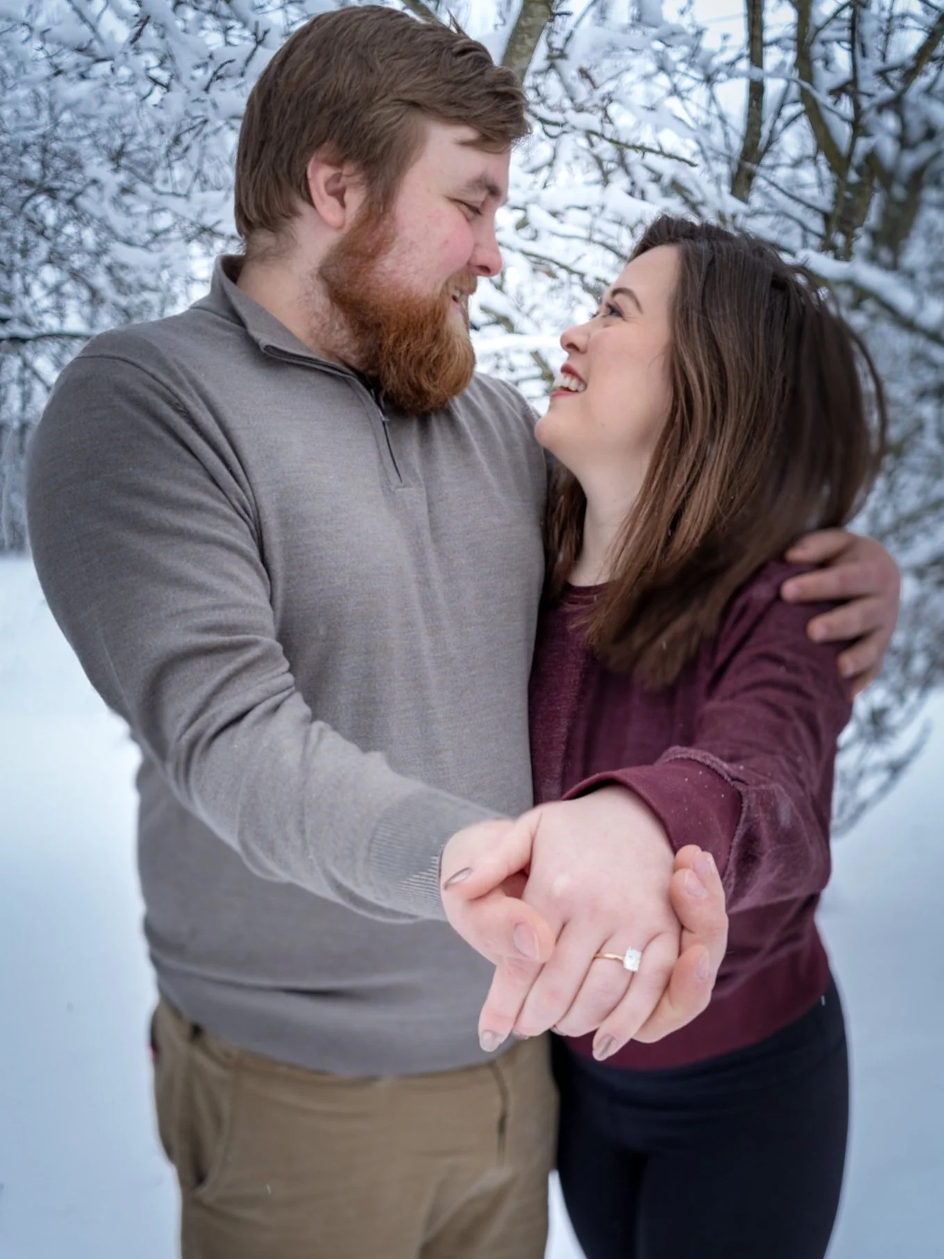 A couple is holding hands, standing outside in a snowy winter landscape, looking at each other affectionately.
