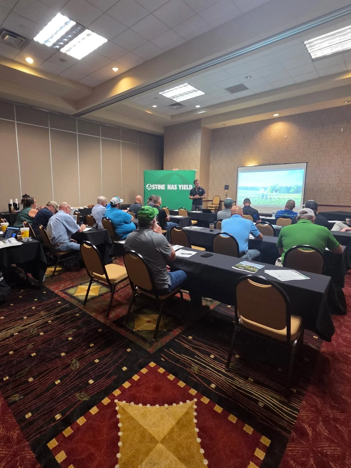 A group of people attending a presentation  in a conference room with a speaker from Stine Seed Company at the front and a large screen displaying a scenic image. The room has patterned carpet and tables with black tablecloths.
