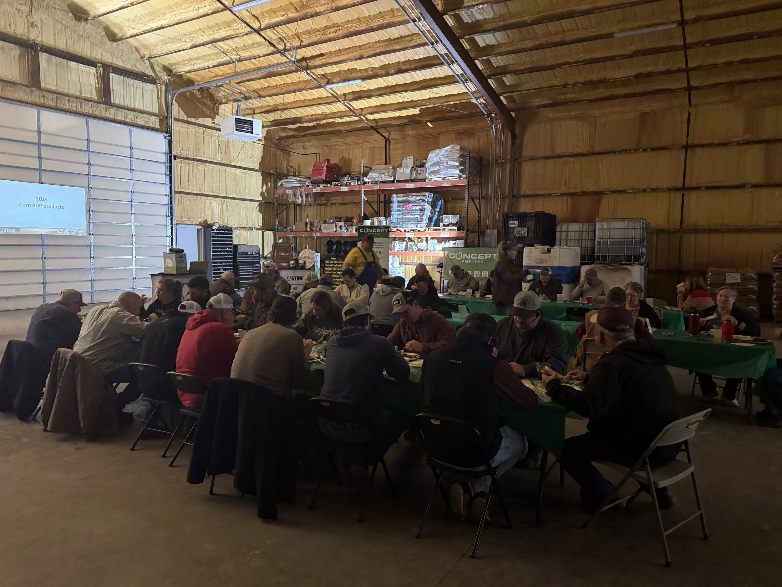 People sitting at tables inside a large warehouse or barn, with a high ceiling and metal and wooden walls, attending an event or meeting.