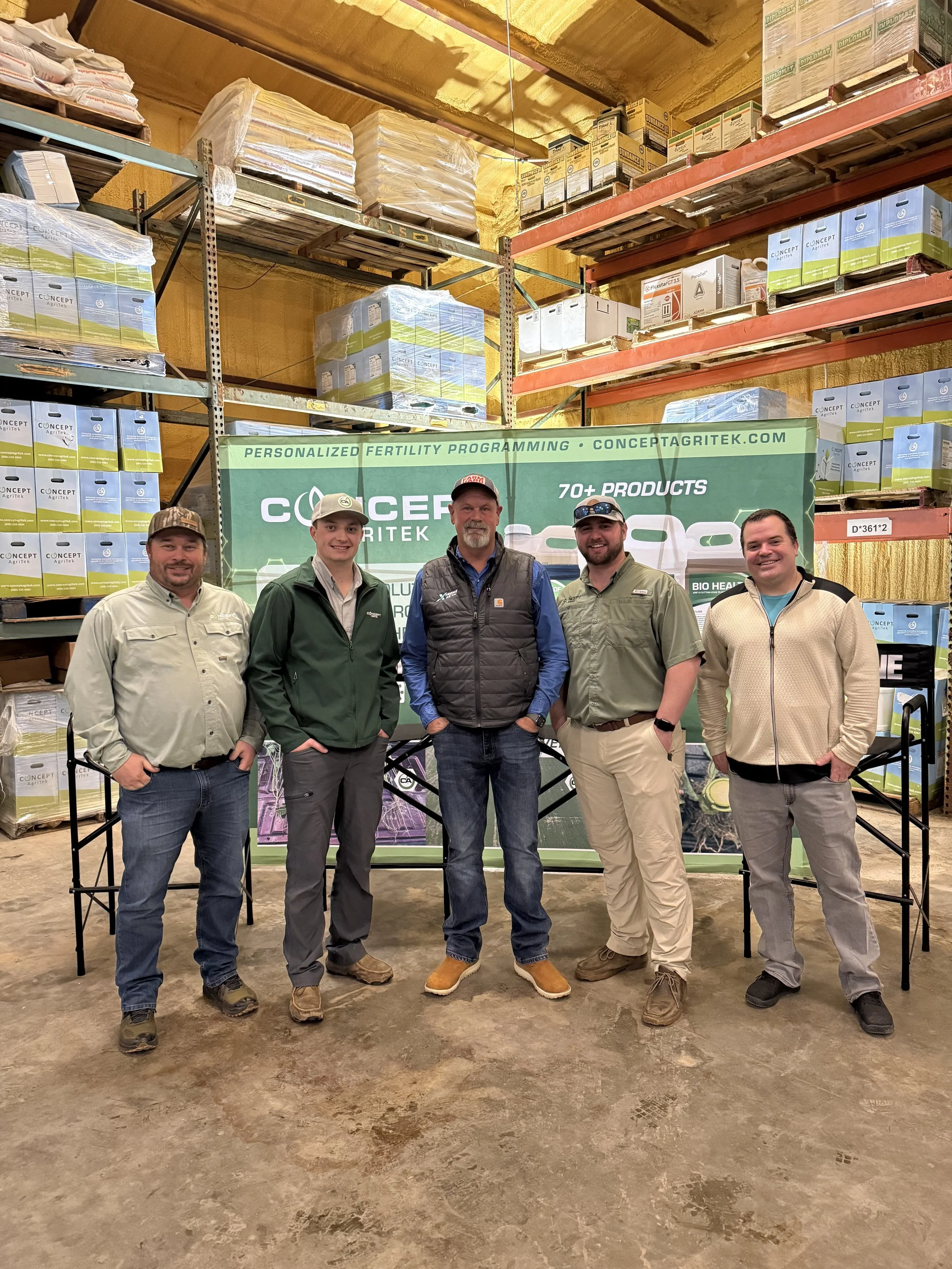 Five men standing in front of a green and white promotional display and warehouse shelves filled with boxes and bags of agricultural products.