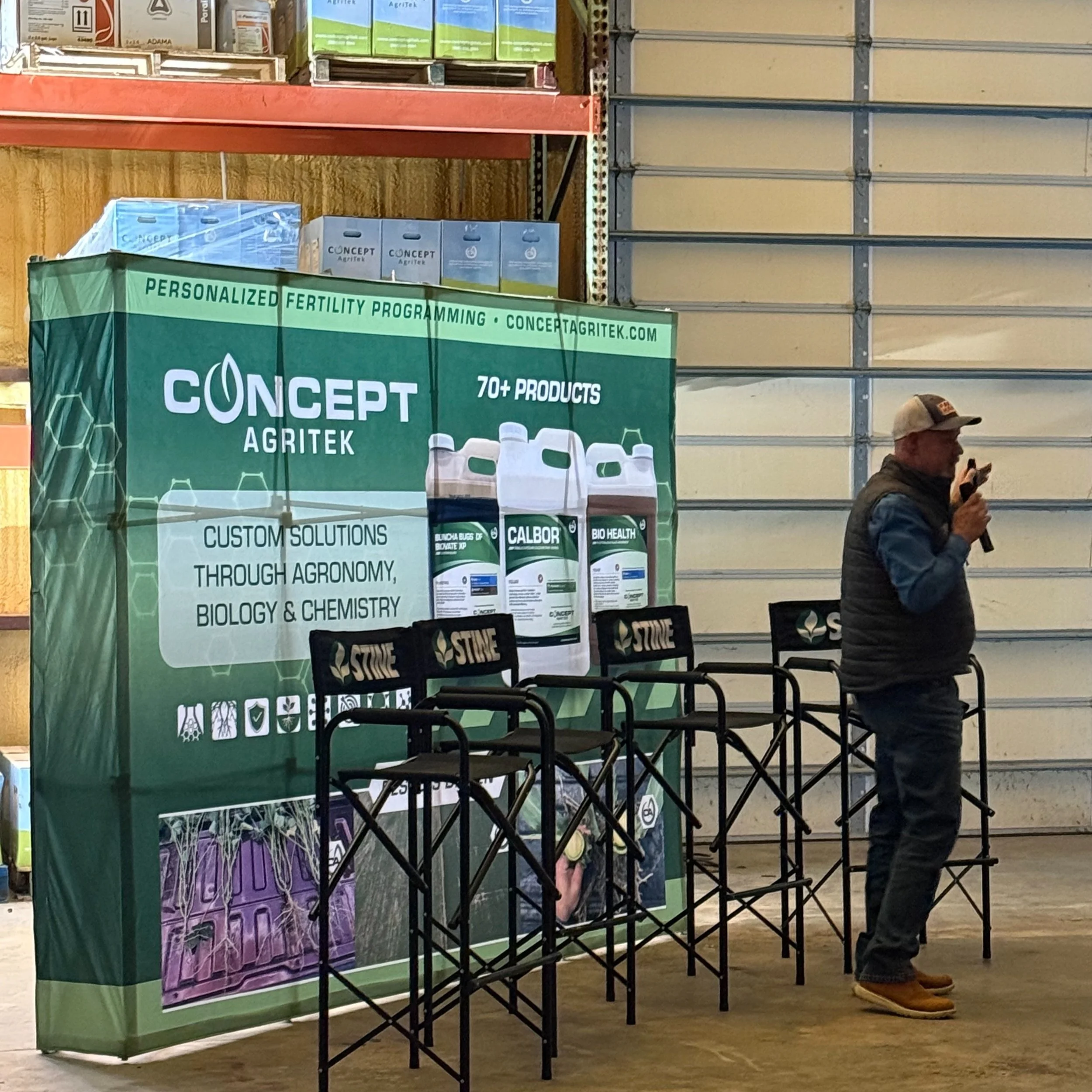 At a store display, a man wearing a baseball cap, glasses, a dark jacket, and brown shoes is looking at a cell phone. An upright banner features the brand 'Concept Agritek' with green and purple packaging images, promoting personalized fertility programs, agritech solutions, and biochemistry products, on a wooden floor with chairs in front.