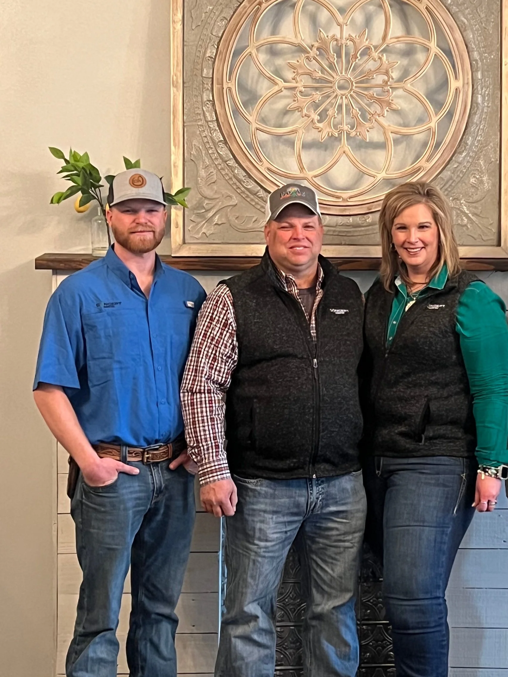 Three people standing indoors in front of a decorative wall hanging with a large circular design and glass window. The person on the left is a man in a blue shirt and gray cap, the middle person is a man in a checkered shirt and black vest, and the right person is a woman with blonde hair wearing a green and black jacket.