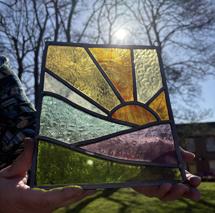 Colourful stained glass panel with a sun design, held up against the sunlight outdoors.