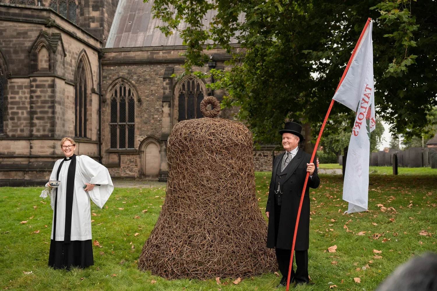 A woman dressed as a clergyperson, a man dressed in Victorian-style clothing with a top hat, and a large woven sculpture of a bell are outside a historic church. The man holds a white flag, and the woman is holding a cup and a small tray. The scene takes place on a grassy area with autumn leaves and trees, with a stone church in the background.