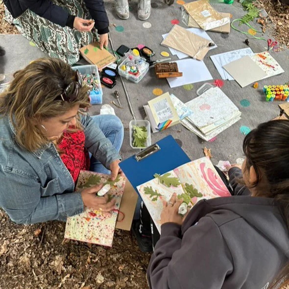 Two women making crafts on a sidewalk, with craft supplies, paper, and decorations scattered around them.