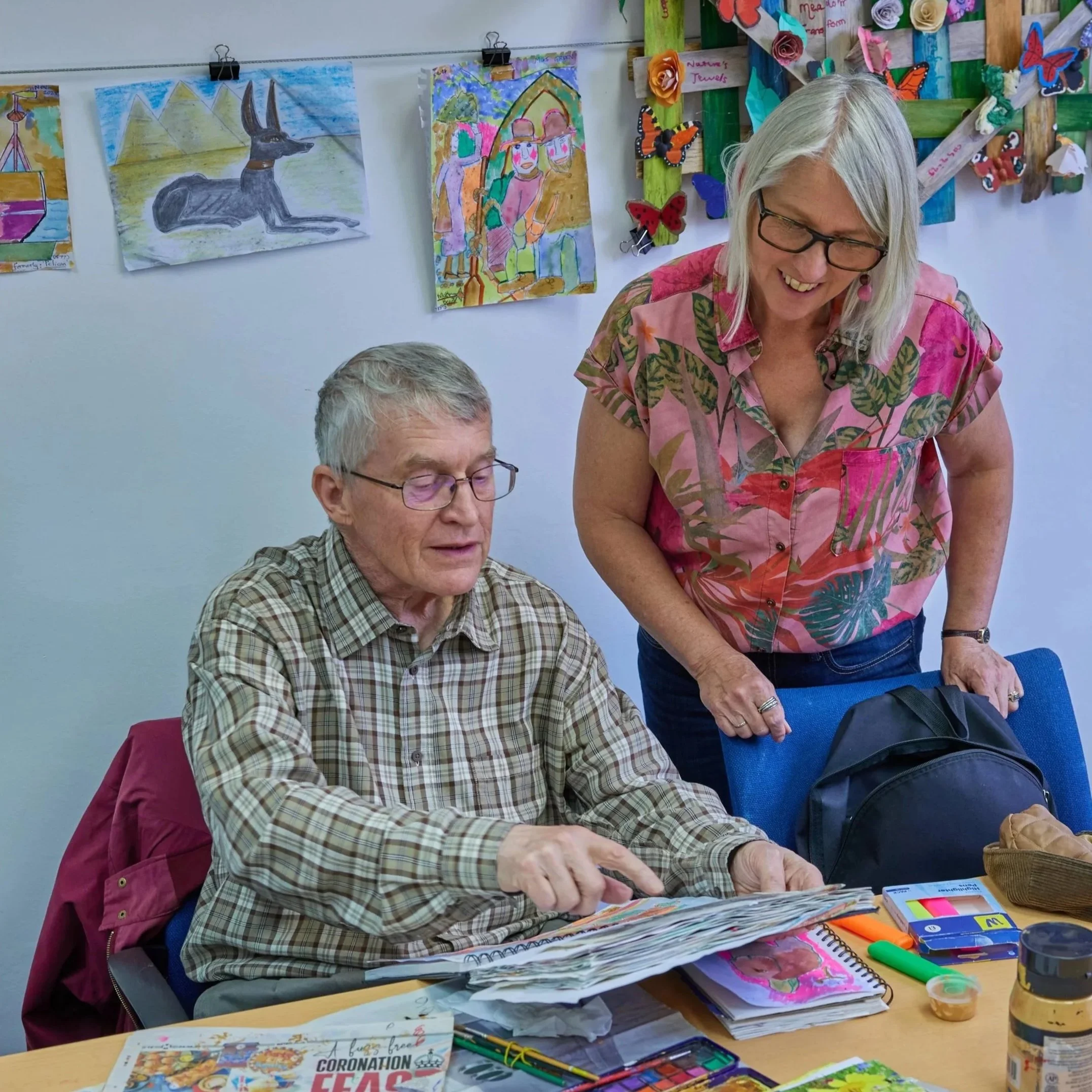 A Charnwood Arts Drawing Room session with a man with glasses and a striped shirt is sitting at a table, looking at a sketchbook with a woman wearing a colorful pink and green floral shirt, stands next to him, smiling and leaning over the table. 