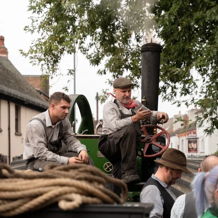Three men sitting on a vintage green tractor, one is reading a mobile phone, and the others are looking away or down, outdoors with trees and buildings in the background.