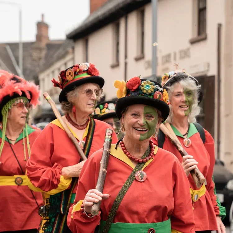A group of elderly women dressed in colorful costumes, carrying sticks, and wearing decorated hats, participating in a street parade or festival.