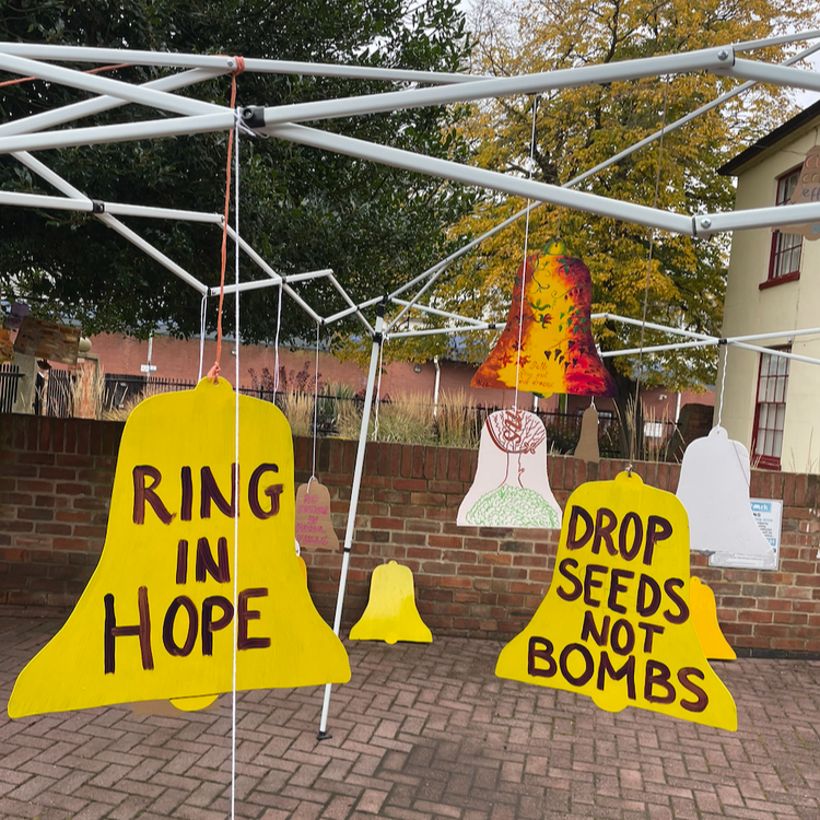 Hanging signs in a garden display with messages like 'Ring in Hope' and 'Drop Seeds Not Bombs', with a brick wall and trees in the background.