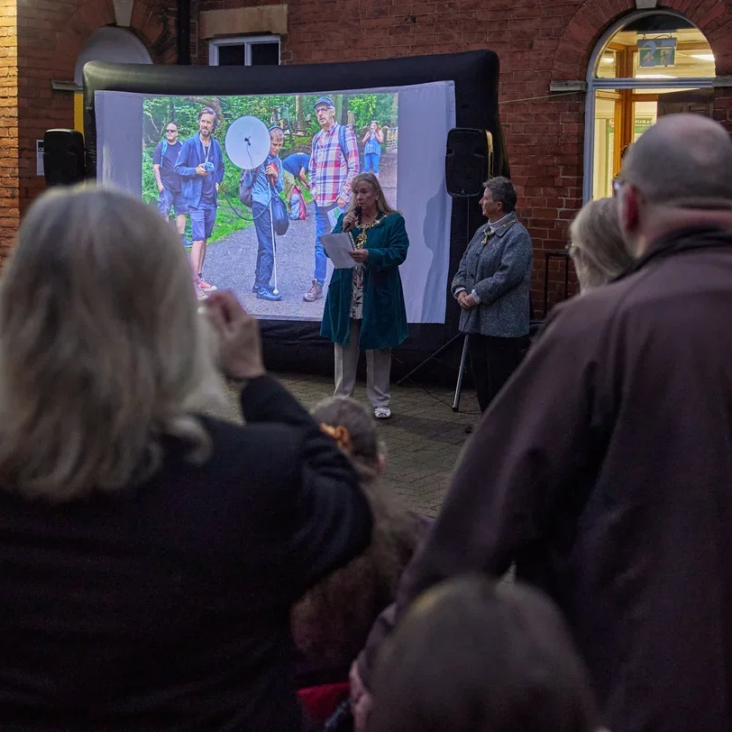 Shirley Novak speaking at the Loughborough Bell Festival 2024. An outdoor event, in front of a large outdoor screen displaying a photo of several people, with several people in the audience listening.