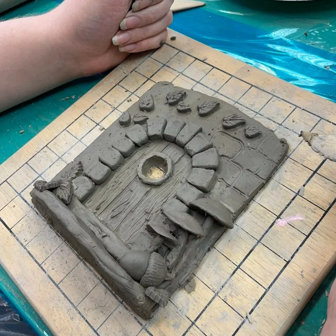 Clay sculpture of a wooden door with a circular handle, surrounded by a stone archway and walls, on a square base with a grid pattern.