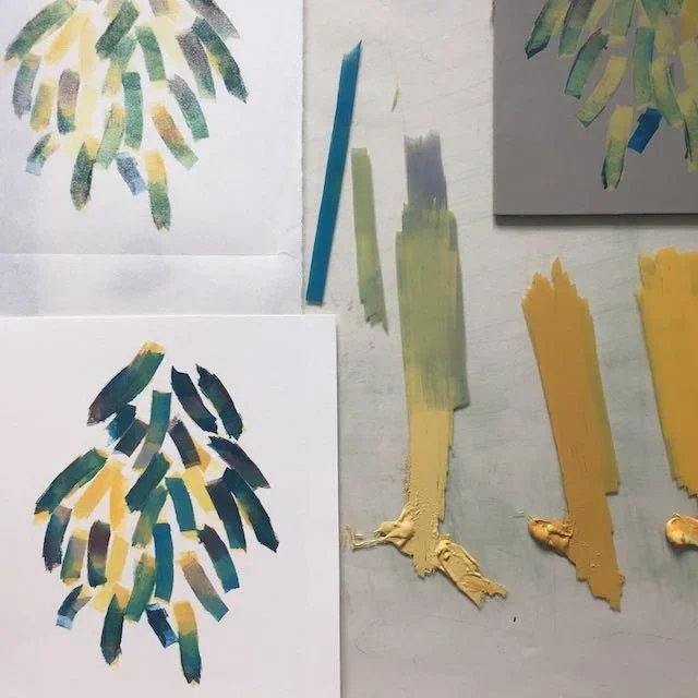 Two painted paper art pieces with abstract leaf patterns, along with various paint swatches, brushes, and paint tubes on a table.