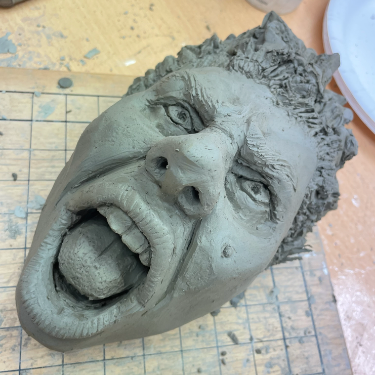 Clay sculpture of a person's head with an open mouth, detailed facial features, and curly hair, on a work surface.