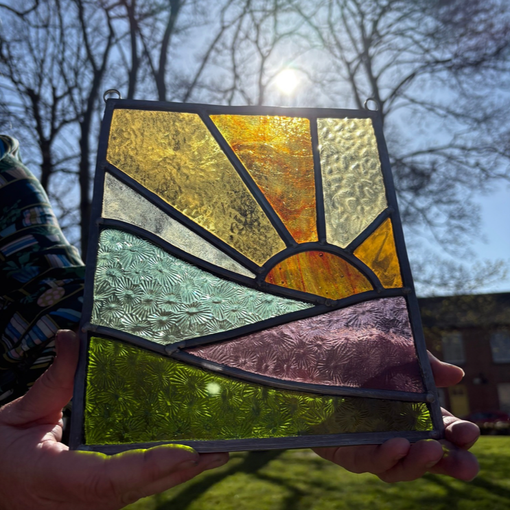 A stained glass panel depicting a sunrise or sunset with colorful glass sections, outdoors on a sunny day with trees and buildings in the background.