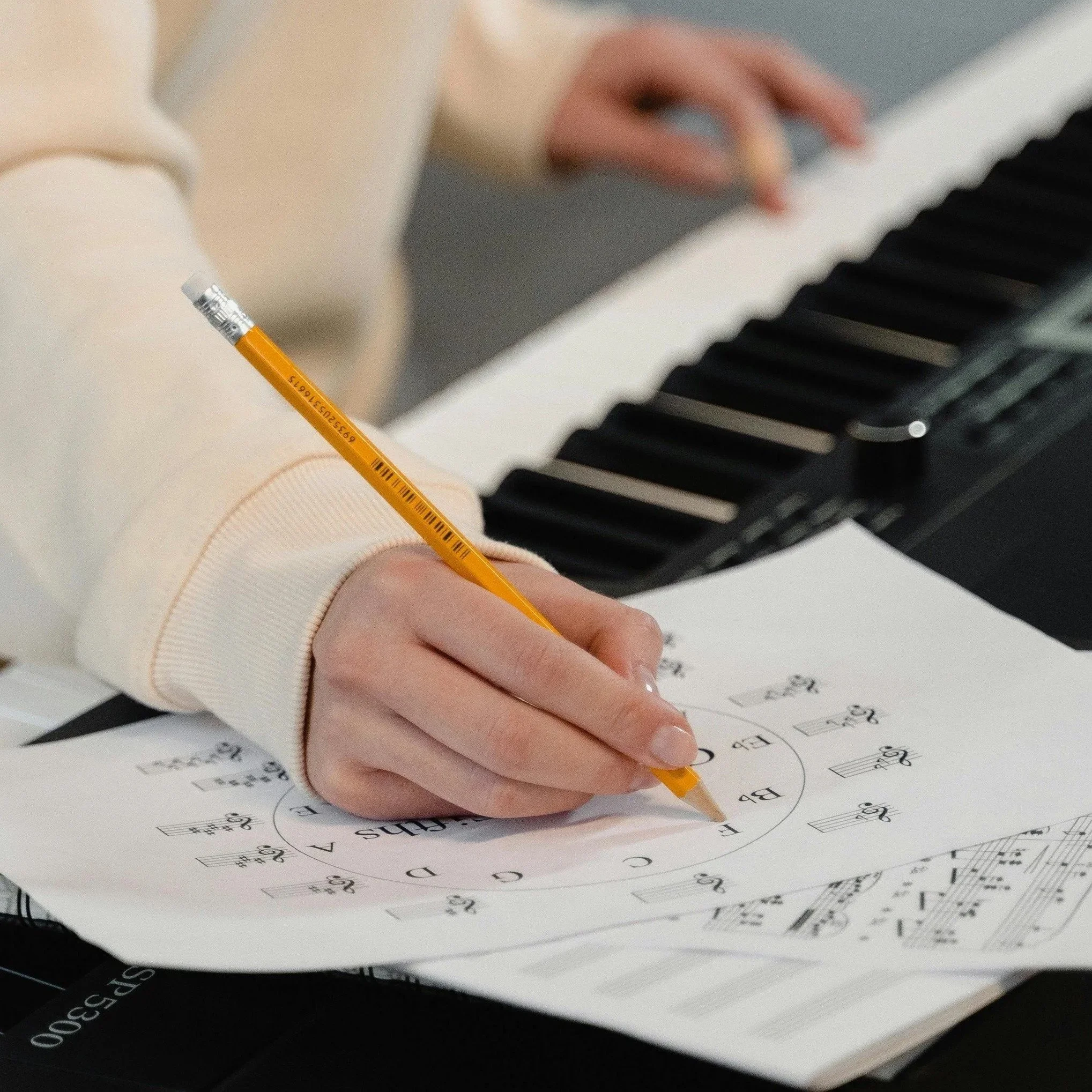 A person writing musical notes and symbols on sheet music with a yellow pencil, with a digital keyboard in the background.