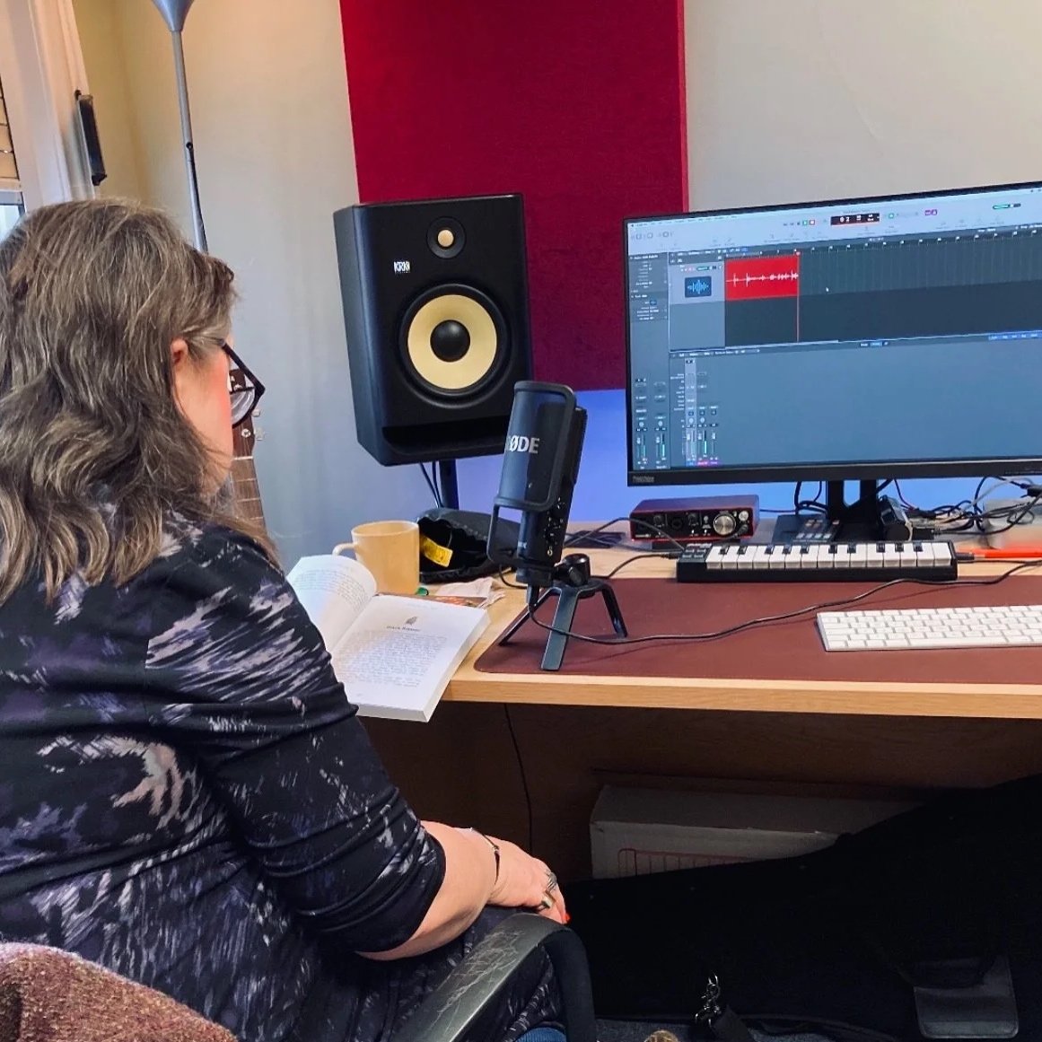 Woman recording music in a studio with a computer, speaker, microphone, and open book on the desk.