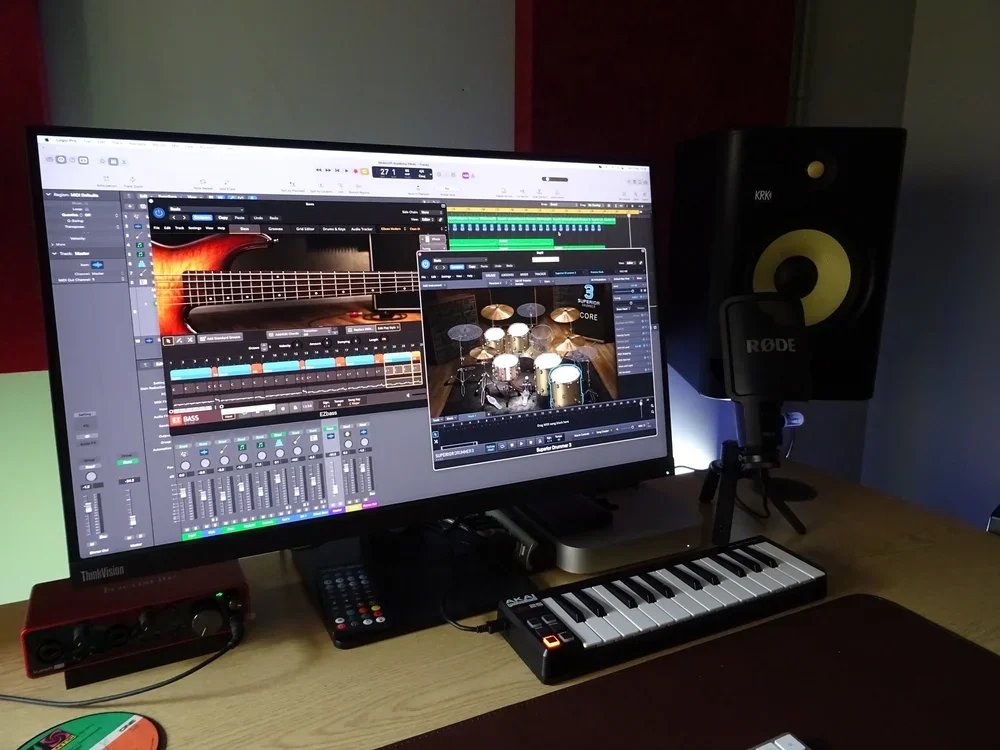 Music production setup with a large monitor displaying a digital audio workstation, a MIDI keyboard, studio monitor speakers, a Rode microphone, and various music equipment.
