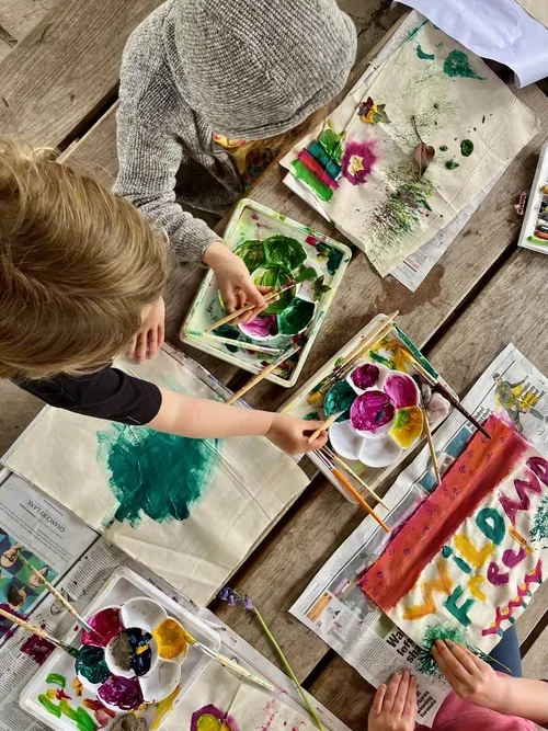 Two children making colourful finger paintings with brushes and finger paints on paper and newspaper, on a wooden table.