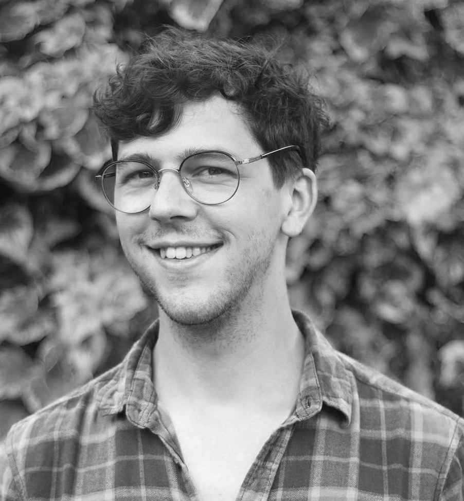 A photo of Michael Arrowsmith with curly brown hair, wearing glasses, smiling, and dressed in a plaid shirt, standing outdoors with green foliage in the background.