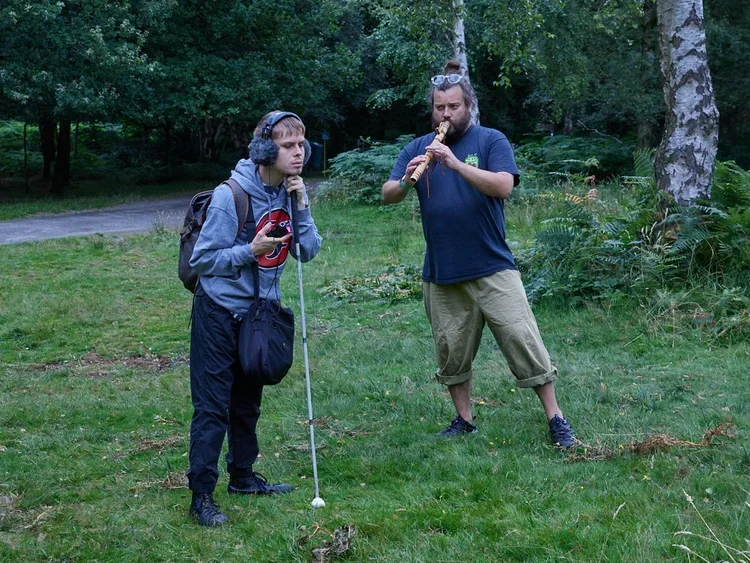 A man with a backpack and headphones listening to a man playing a flute outdoors in a forest.