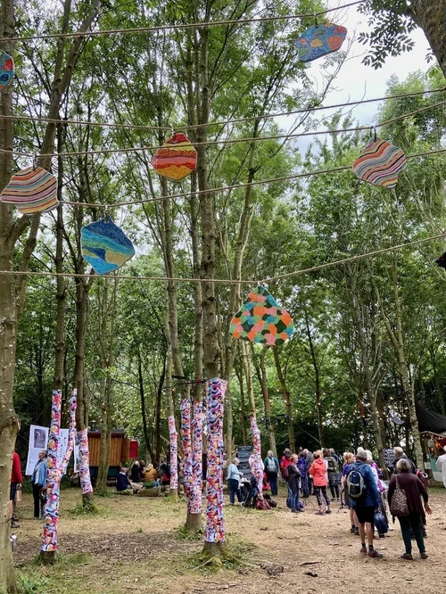 Outdoor festival with colourful hanging art and people gathered among trees.