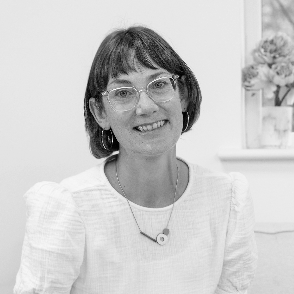 A photo of Lucy Bartram with short brown hair, glasses, and hoop earrings smiling at the camera, sitting indoors with a window and pink flowers in the background.