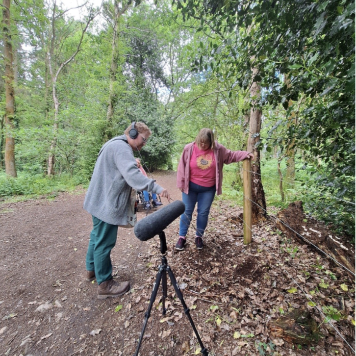 Two people recording audio in a wooded outdoor area, with a boom microphone and headphones, on a trail surrounded by trees and green foliage.