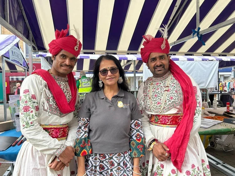Three people, two men in traditional Indian attire with red turbans and a woman in casual clothing, standing under a striped canopy at an outdoor event or festival.