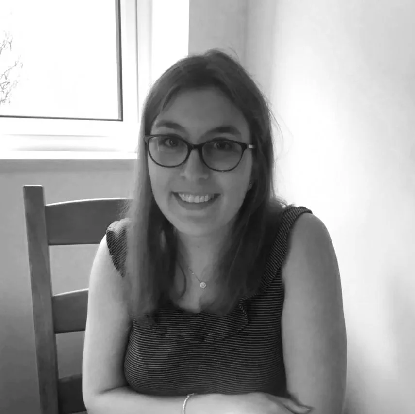 A photo of Charlotte Curran with glasses and long brown hair smiling at the camera, seated at a wooden table near a window in a well-lit room.